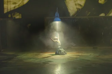 A performer kneels on the empty stage directly beneath a large, hanging lamp. He is illuminated by the harsh light and gazes into it.