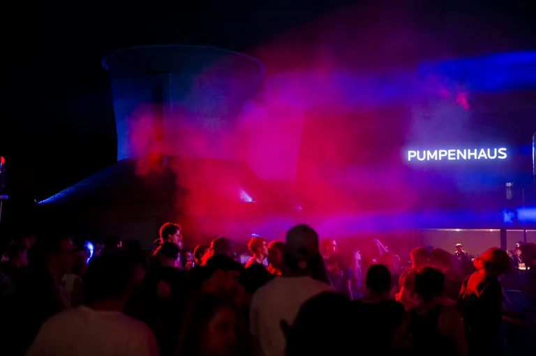 A nighttime scene at a party with many people dancing in colorful lights. Red and blue lights, along with fog, fill the air, creating an atmospheric mood. In the background, a sign with the word "PUMPENHAUS" is glowing.