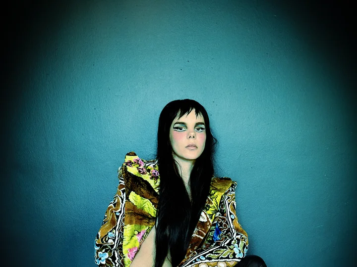 Gaye Su Akyol hockt vor einer blaugrünen Wand. Sie hat lange, schwarze Haare und trägt auffälliges Make-up, eine gemusterte Jacke mit üppigen Schultern und hochhackige Lederstiefel. Unter ihr liegt ein Tuch mit einem floralen Muster. Sie blickt direkt in die Kamera.