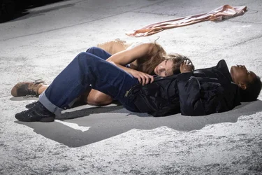 Two performers lie close together on a light-colored, sloping stage surface. One wears dark clothing and jeans, while the other wears a light-colored costume and rests her head on the other performer’s shoulder. A long strip of pink fabric lies in the background.
