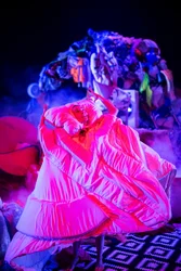 Performer:in auf der Bühne, vollständig in eine große, pink angeleuchtete Bettdecke gehüllt; im Hintergrund ein hoher Stapel aus Kleidung und Alltagsobjekten.