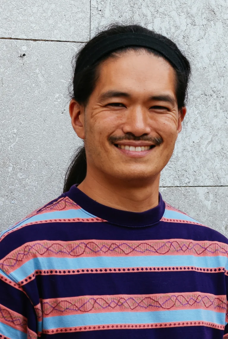Takeshi Matsumoto smiles at the camera. He is wearing a colorful, patterned, striped shirt in shades of blue, pink, and purple.