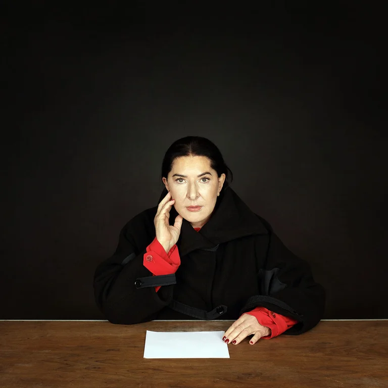 Portrait of Marina Abramović in a black coat with red sleeves, seated at a table and looking straight at the camera.