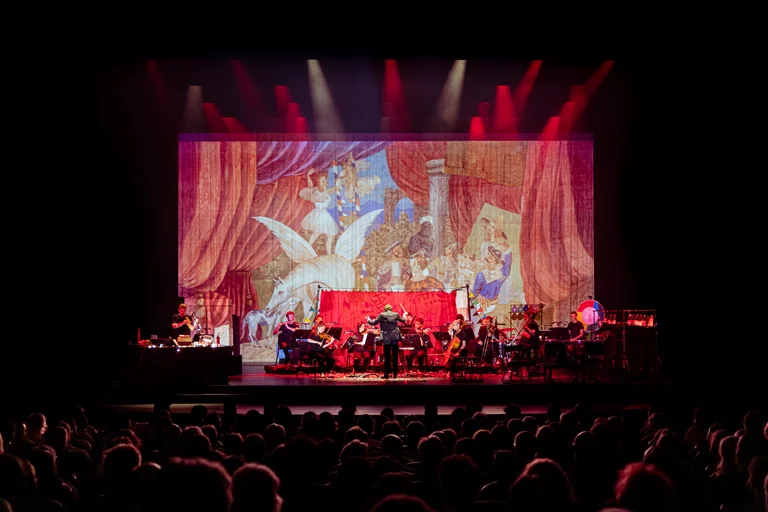 The ensemble Les Apaches ! performs on a large stage in front of an audience under red stage lighting. In the background, a large-scale projection shows a colorful, theatrical scene.