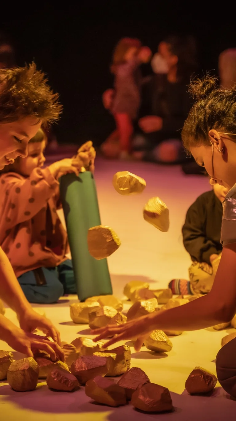 Szene aus der partizipativen Performance UNIVERS: Performer:innen und Kinder sitzen auf dem Boden und spielen gemeinsam mit kleinen, steinähnlichen Objekten.