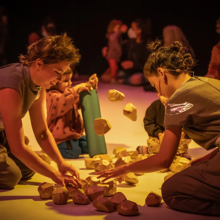 Scene from the participatory performance UNIVERS: Performers and children sit on the floor, playing together with small stone-like objects.