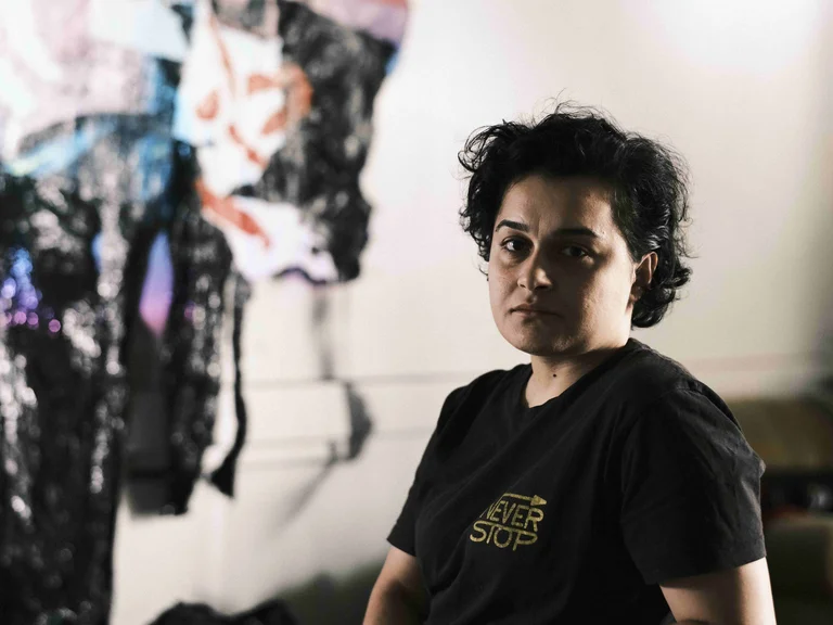 Hülya Özkan sits at a table in a studio space, looking at the camera; behind her hangs an abstract, colorful wall piece.