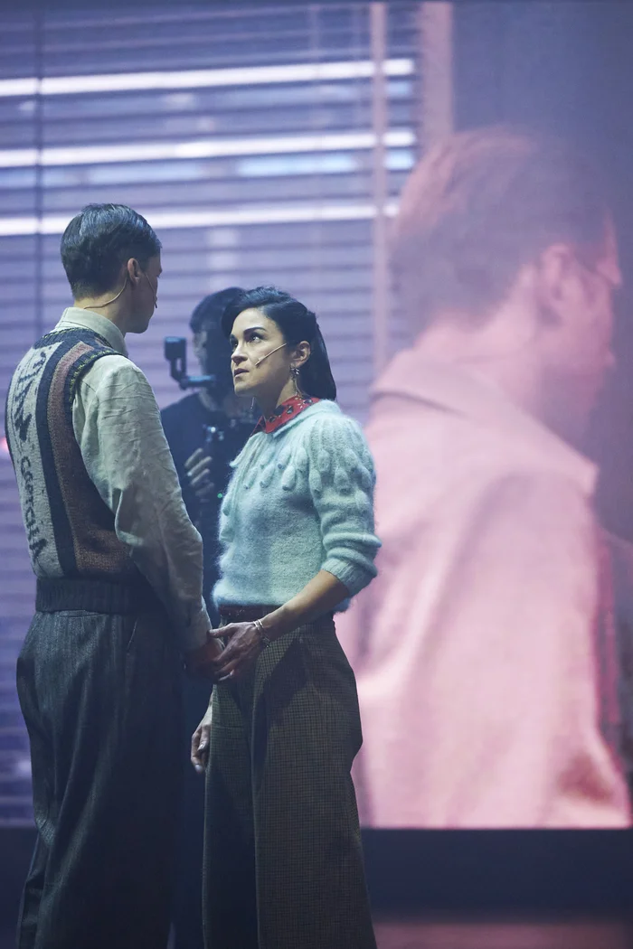 Two actors face each other on stage, holding hands, with a projection of their faces in the background.