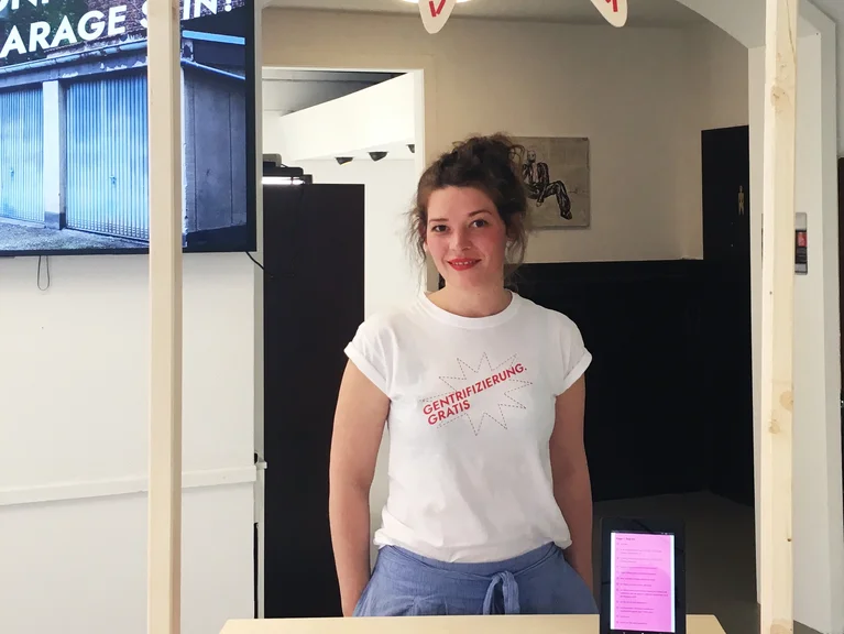 A person stands at an information booth beneath a sign reading "Gentrifizierung gratis" ("Gentrification for free").