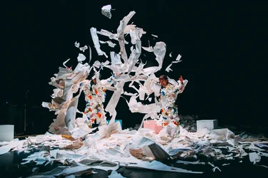 Die Performer:innen Takeshi Matsumoto und Makiko Aoyama in Kostümen aus buntem Papier werfen Papierfetzen in die Luft, umgeben von verstreuten Blättern.