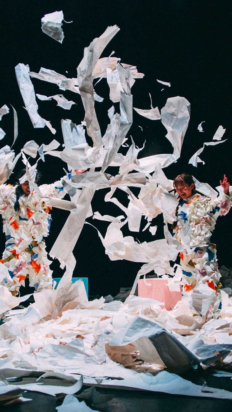 Die Performer:innen Takeshi Matsumoto und Makiko Aoyama in Kostümen aus buntem Papier werfen Papierfetzen in die Luft, umgeben von verstreuten Blättern.