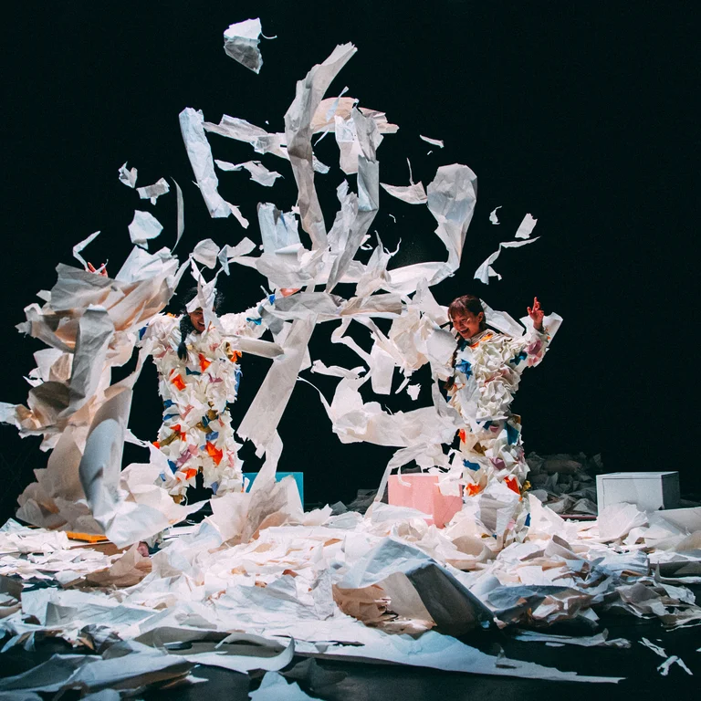 The performers Takeshi Matsumoto and Makiko Aoyama in colorful paper costumes throw paper fragments into the air, surrounded by scattered sheets.