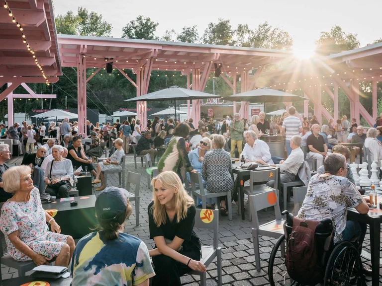 Ein lebhaftes Publikum unter Sonnenschirmen und der rosa Holzkonstruktion des Festivalzentrums Wunderland, im Hintergrund sind Veranstaltungsstände sichtbar.