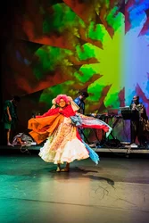 Eine Tänzerin in einem bunten, geschichteten Kleid mit ausladenden Ärmeln tanzt auf einer Bühne. Im Hintergrund sind Musiker an ihren Instrumenten und ein leuchtendes, kaleidoskopartiges Bühnenbild zu sehen.