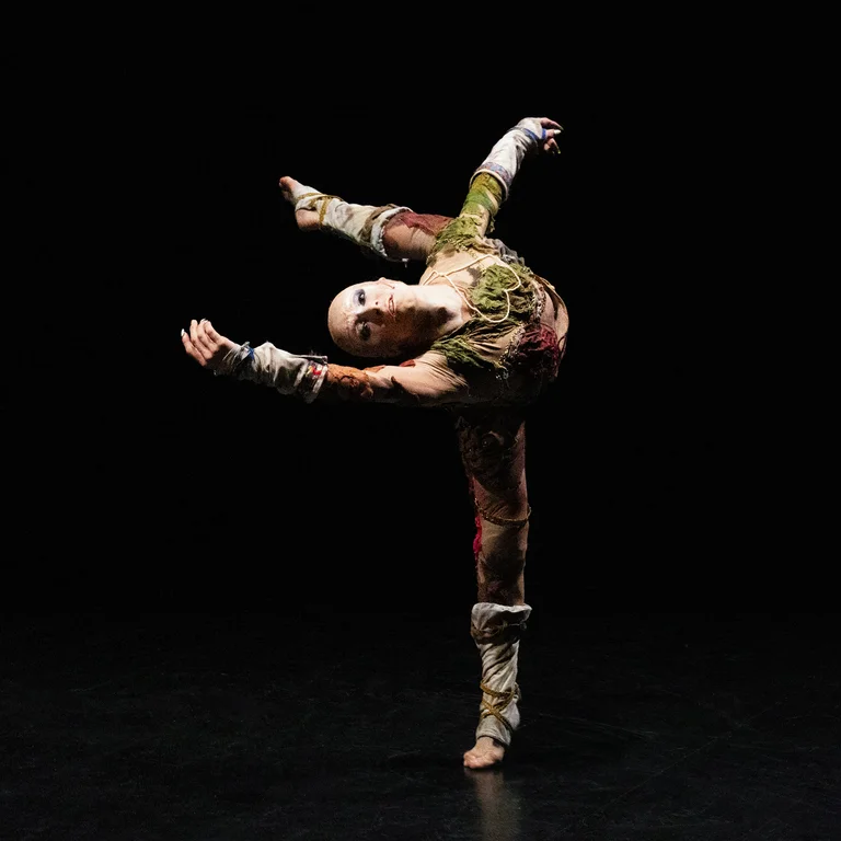 Eine Performerin führt auf einer dunklen Bühne eine dynamische Tanzpose mit ausgestreckten Armen und Beinen im Gleichgewicht aus.