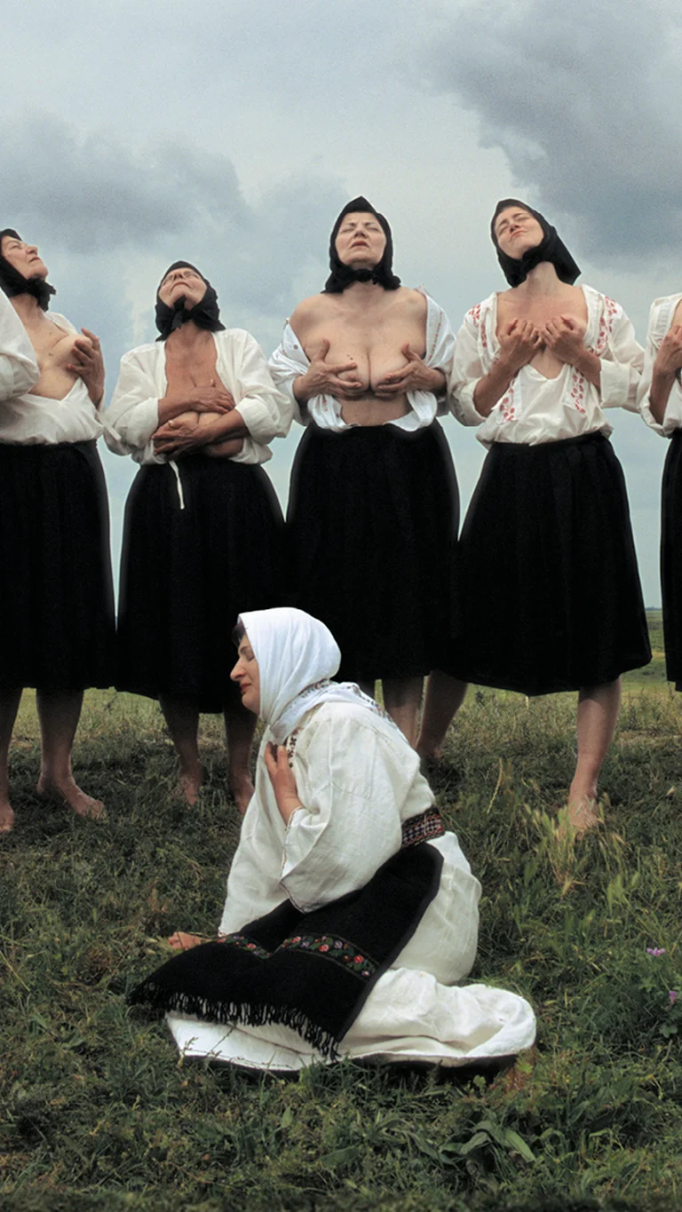 Scene from Balkan Erotic Epic: A group of women stands barefoot in a circle on a field, lifting their heads toward the sky and holding their uncovered chests. Another woman kneels in traditional clothing in the center.