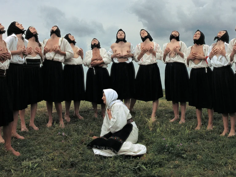 Szene aus Balkan Erotic Epic: Eine Gruppe von Frauen steht barfuß im Kreis auf einer Wiese, hebt den Kopf zum Himmel und hält ihre entblößten Oberkörper. Eine weitere Frau kniet in traditioneller Kleidung in der Mitte.