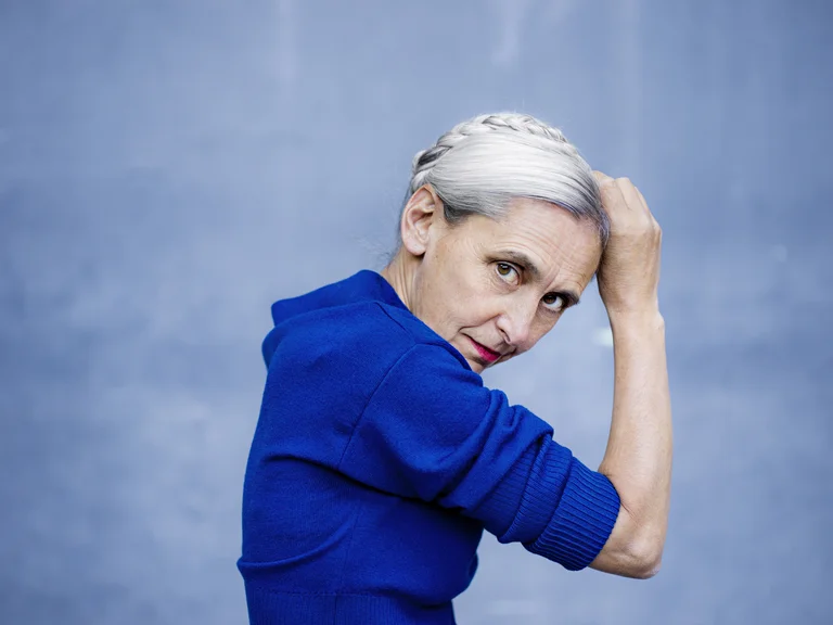 Blonde Frau mit Dutt und blauem Oberteil, seitlich in die Kamera blickend