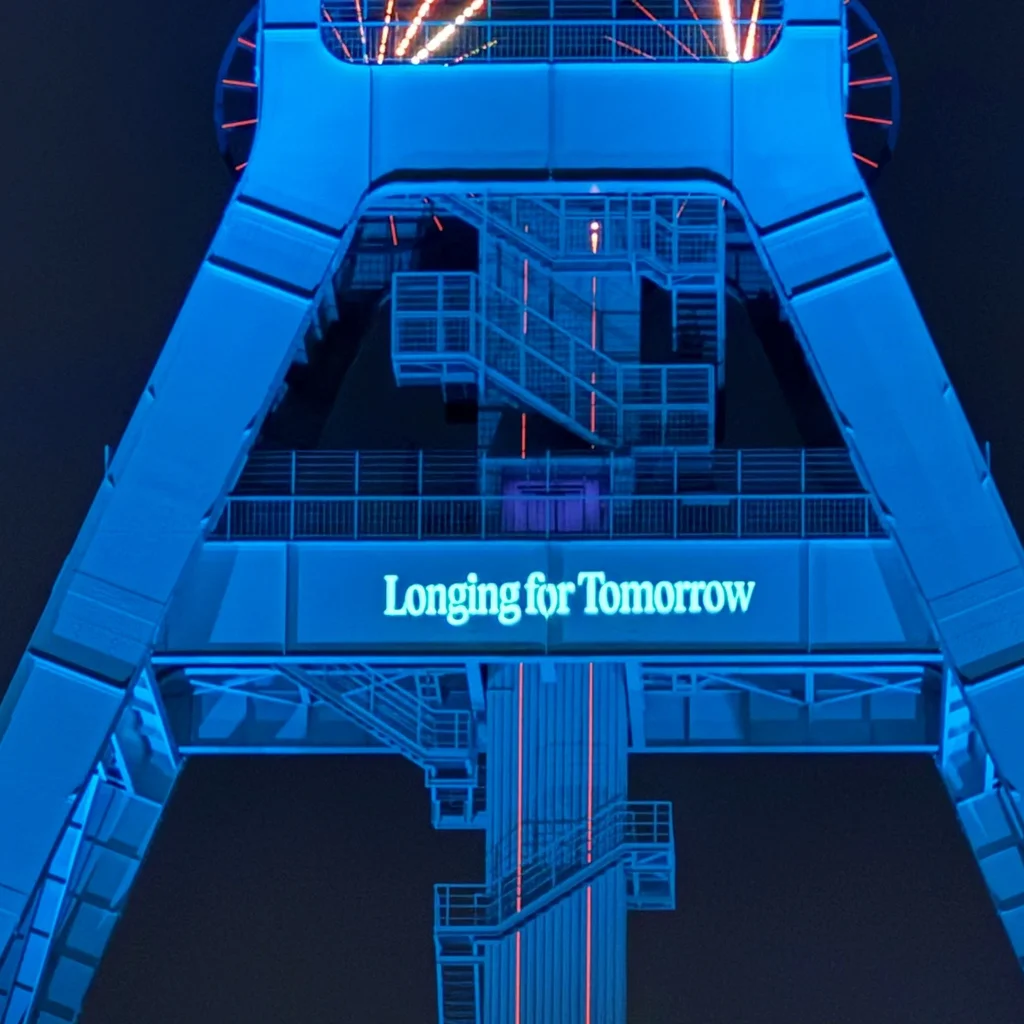 Blau beleuchteter Förderturm des Bergbau-Museum Bochum vor dunklem Himmel, mittig mit der leuchtenden Schrift „Longing for Tomorrow“