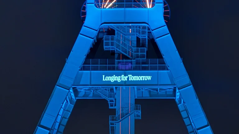 Blau beleuchteter Förderturm des Bergbau-Museum Bochum vor dunklem Himmel, mittig mit der leuchtenden Schrift „Longing for Tomorrow“