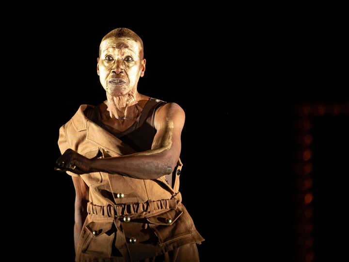 Germaine Acogny mit goldener Farbe in Gesicht und auf ihrem linken Arm, den sie gebeugt vor ihren Körper hält.