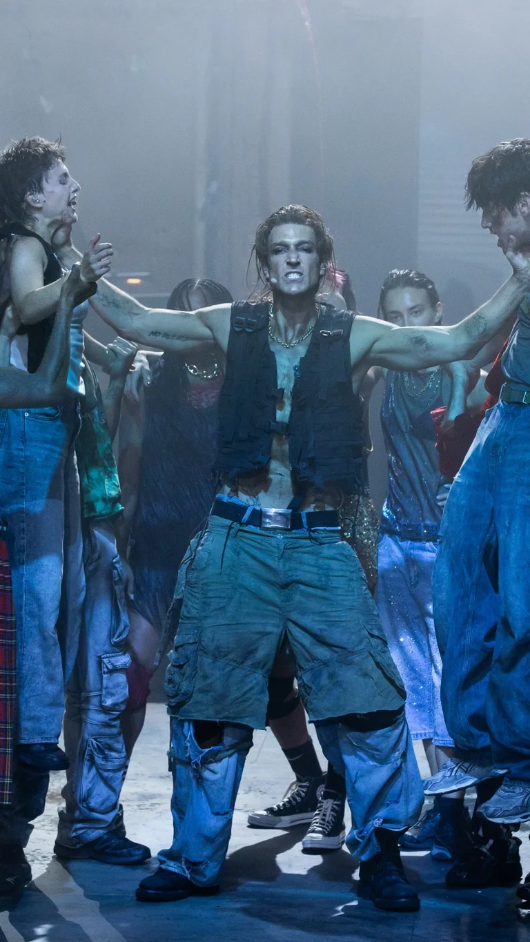 The cast of Rebel Rebel on a stage bathed in cold light. In the centre is the actor Daniel Donskoy, who is holding two other performers by the neck and lifting them into the air.