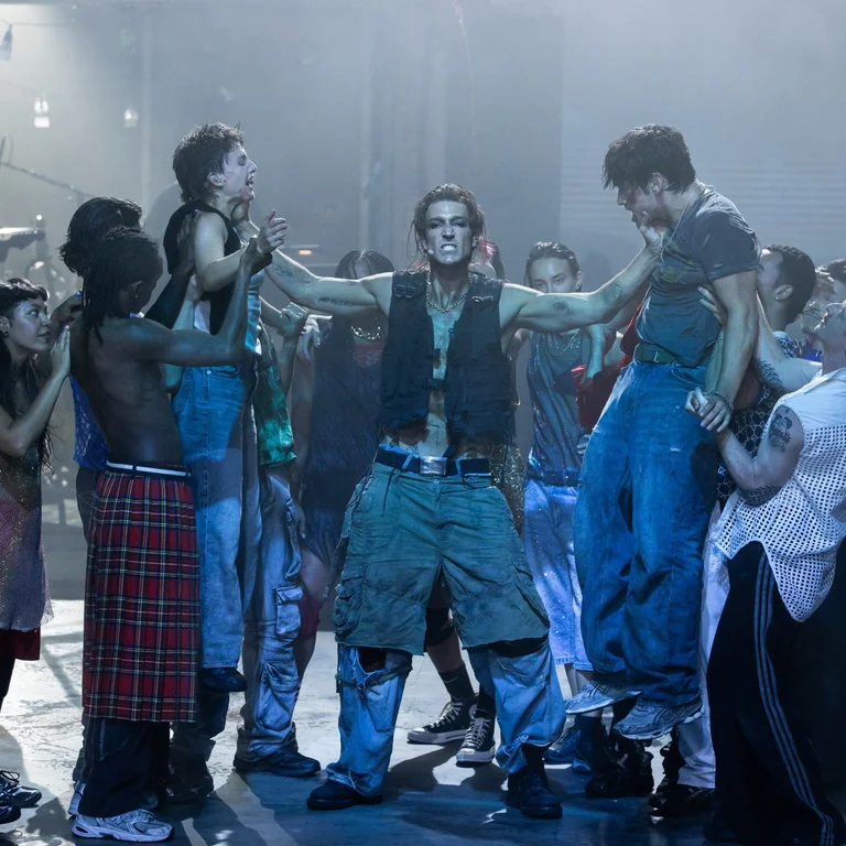 Der Cast von Rebel Rebel auf einer in kaltem Licht ausgeleuchteten Bühne. In der Mitte der Schauspieler Daniel Donskoy, der zwei weitere Darstellende am Hals hält und in die Luft hebt.