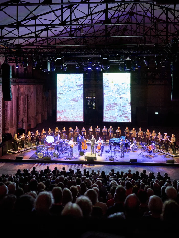 Conductor Florian Helgath with Chorwerk Ruhr and Bang on a Can All-Stars on stage, audience in the foreground, with two projections of abstract light blue patterns in the background.