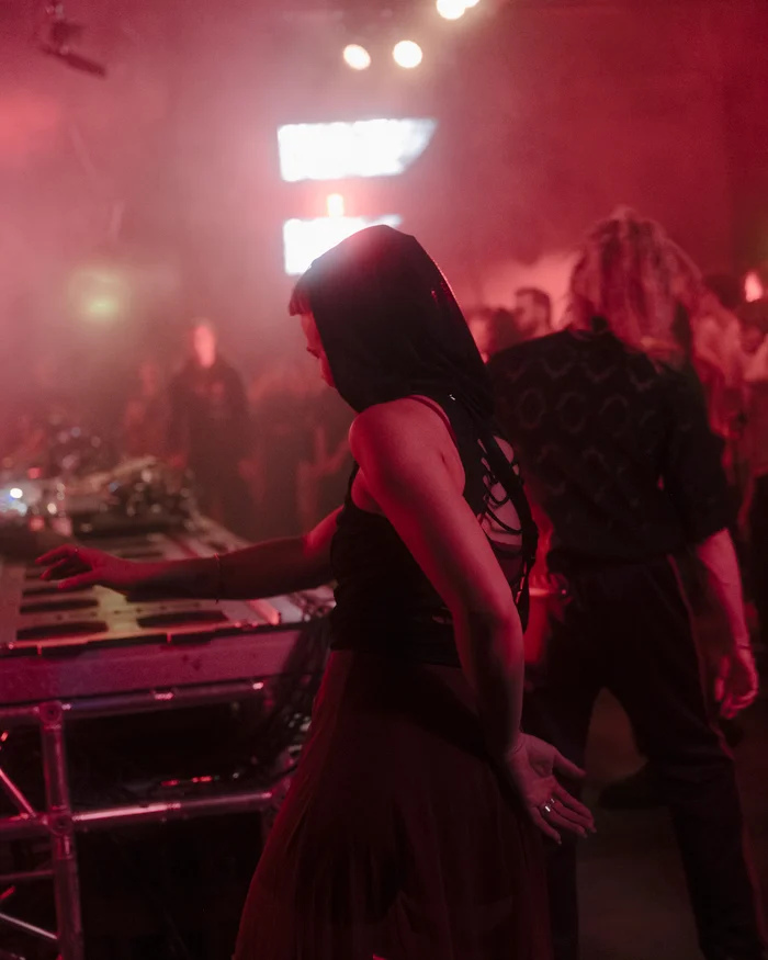 A person wearing a hood dances in red light next to a DJ booth, surrounded by other people.