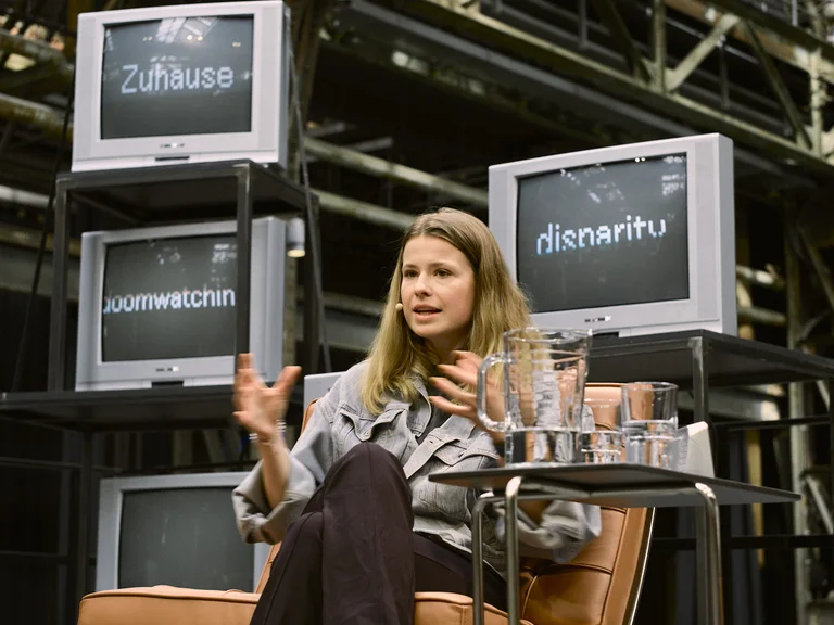 Luisa Neubauer speaks on stage at the event Brave New Voices and gestures during her contribution. She sits in a leather chair next to a table with water glasses and a carafe. In the background, several old television sets are visible, displaying words such as ‘Zuhause’ and ‘disparity’ on their screens.