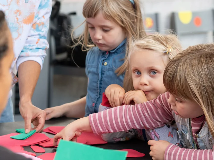 Mehrere Kinder sitzen an einem Tisch und greifen nach bunten Papierformen, während eine erwachsene Person den Workshop anleitet.
