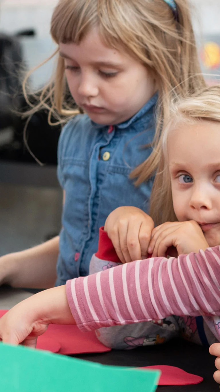 Mehrere Kinder sitzen an einem Tisch und greifen nach bunten Papierformen, während eine erwachsene Person den Workshop anleitet.