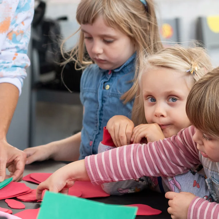 Mehrere Kinder sitzen an einem Tisch und greifen nach bunten Papierformen, während eine erwachsene Person den Workshop anleitet.