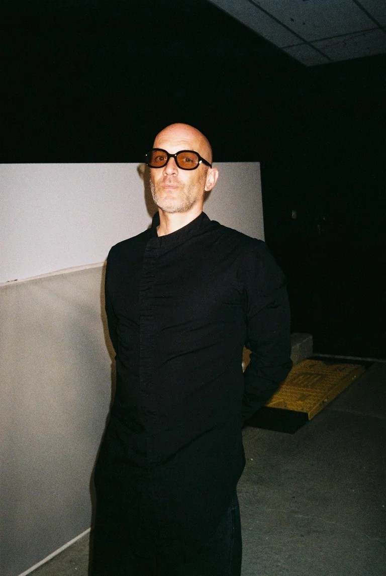 Yvan Talbot stands in a dark room and looks directly into the camera. He is wearing a long black outfit and tinted glasses. His hands are clasped behind his back. In the background, a light-colored wall and a simple platform are visible.