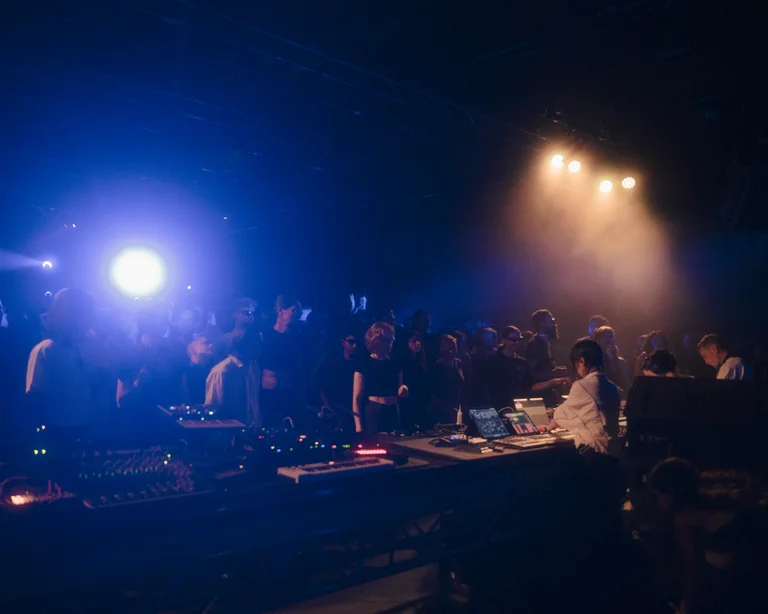 A DJ performs at a mixing desk while a crowd gathers in the blue and orange glow of the stage lights. The intense lighting and haze create a pulsating club atmosphere.