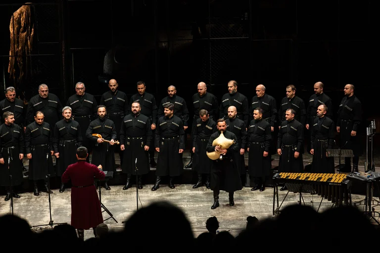 A male choir sings in a hall, in front of them the conductor in a red robe and a bagpiper.