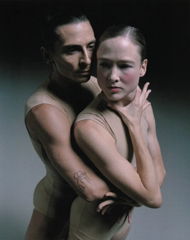 Two dancers in skin-toned costumes stand closely entwined, with one gently holding the other by the neck. The pose conveys both intimacy and tension.