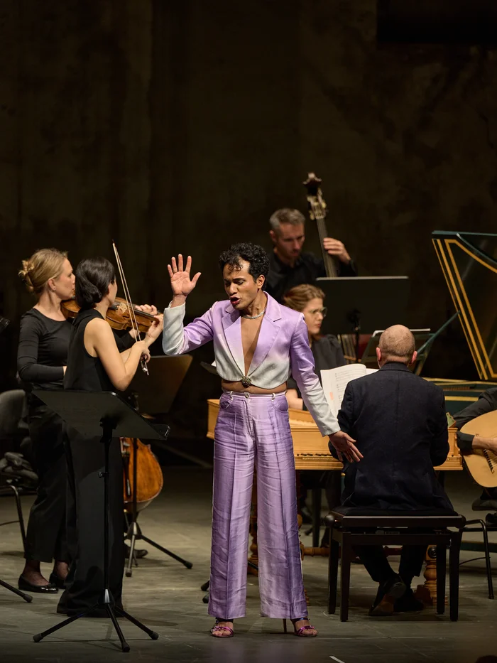Samuel Mariño im lilafarbenen Anzug performt mit erhobener Hand vor einem Barockorchester.