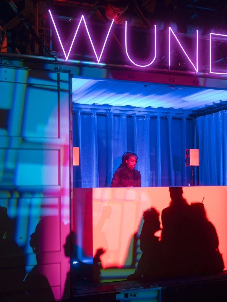 Eine DJ mit Kopfhörern steht in einem blau beleuchteten Container hinter einem rot leuchtenden Pult. Die Schatten tanzender Menschen zeichnen sich auf dem Pult und den Wänden ab, darüber leuchtet der Schriftzug „WUNDER“ in violettem Neonlicht.