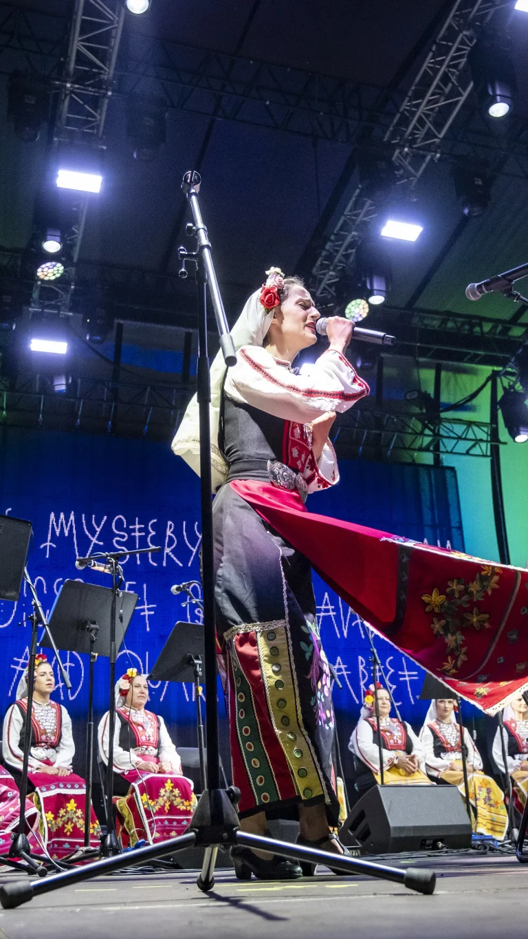 A singer in traditional costume performs into a microphone on a lit stage, while an ensemble of musicians and singers in similar folkloric attire sit behind her.