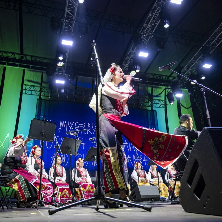 Eine Sängerin in traditioneller Tracht singt auf einer beleuchteten Bühne ins Mikrofon; hinter ihr sitzt ein Ensemble von Musikerinnen und Sängerinnen ebenfalls in folkloristischen Kostümen.