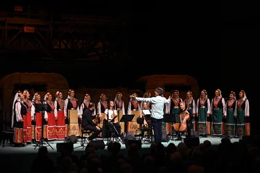Ein Frauenchor in farbenfrohen traditionellen Trachten singt auf einer Bühne, während ein Dirigent mit ausgebreiteten Armen das Ensemble leitet. Vor dem Chor sitzt ein Streichensemble, im Vordergrund ist das Publikum zu sehen.