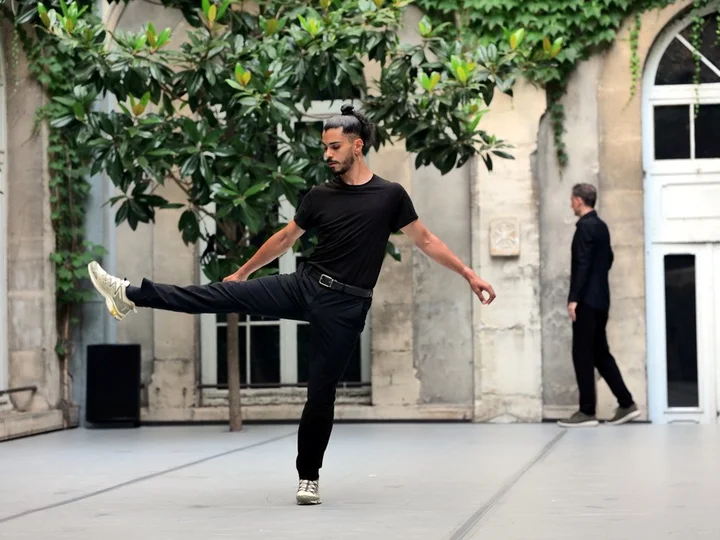Der Tänzer und Choreograf Mohamed Toukabri führt eine ausdrucksstarke Bewegung in einem Innenhof mit grüner, bewachsener Fassade aus. Er trägt ein schwarzes Outfit und hebt ein Bein in die Luft, während er sich mit gestrecktem Arm ausbalanciert. Im Hintergrund ist eine zweite Person in Schwarz zu sehen, die sich in entgegengesetzter Richtung bewegt.