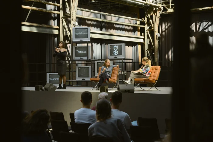 Auf einer Bühne in einer Industriehalle sind drei Frauen Industriehalle. Links steht Konstanze Bustian und dolmetscht in Gebärdensprache. Rechts sitzen Marie Kilg und Sivan Ben Yishai und sprechen miteinander. Hinter ihnen stehen alte Röhrenfernseher, auf denen Begriffe wie „Kapitalismus“, „Thanatos“ und „Staatsräson“ zu sehen sind. Im Vordergrund das Publikum.