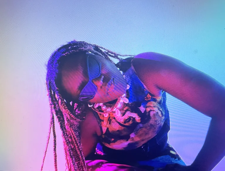 Tatyana Jane wears sunglasses with blue lenses and colorful, patterned clothing. She is bent into a dynamic pose and illuminated by multicolored lighting.