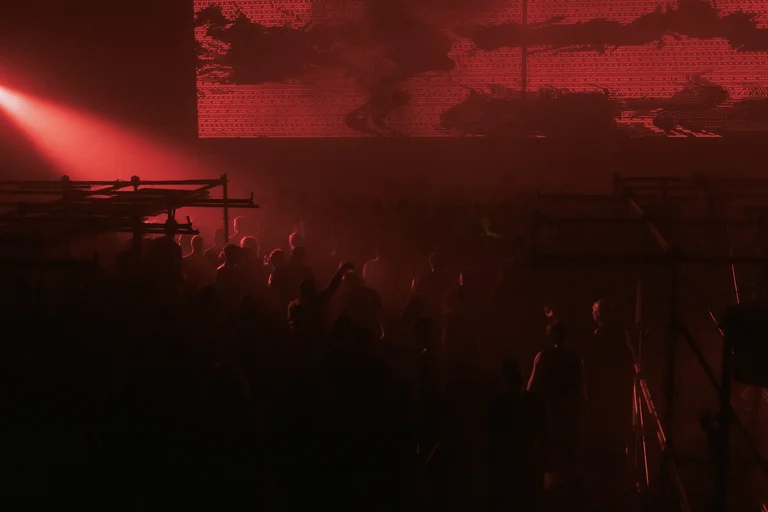 Eine Menschenmenge in einem dunklen, industriellen Raum, beleuchtet von rotem Licht. Im Hintergrund ist eine große LED-Leinwand mit abstrakten, verzerrten roten Mustern zu sehen. Metallgerüste umrahmen die Szene.