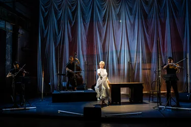 Joyce DiDonato stands on a stage in a white dress next to a wooden table and sings. Around her, the musicians of Time for Three play violin and double bass. Long, brightly lit fabric drapes hang in the background.