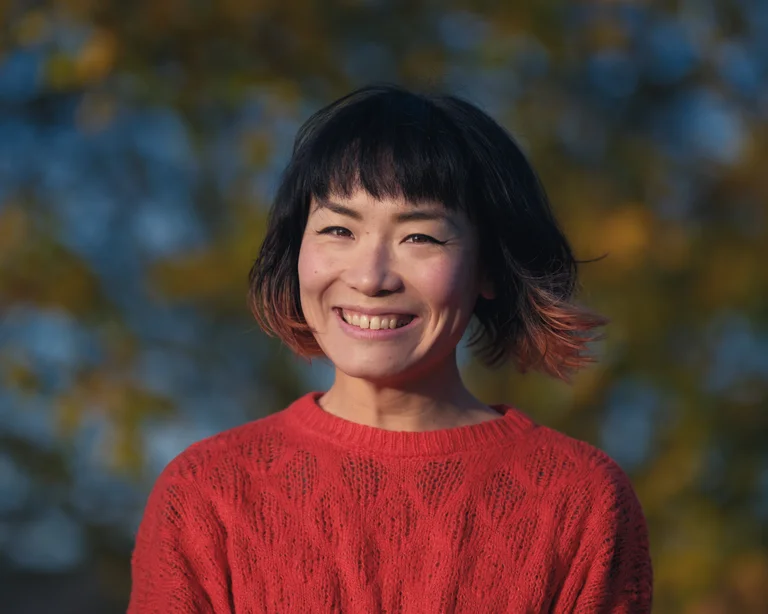 Masumi Saito smiles at the camera. She is outdoors and wearing a red knitted sweater.