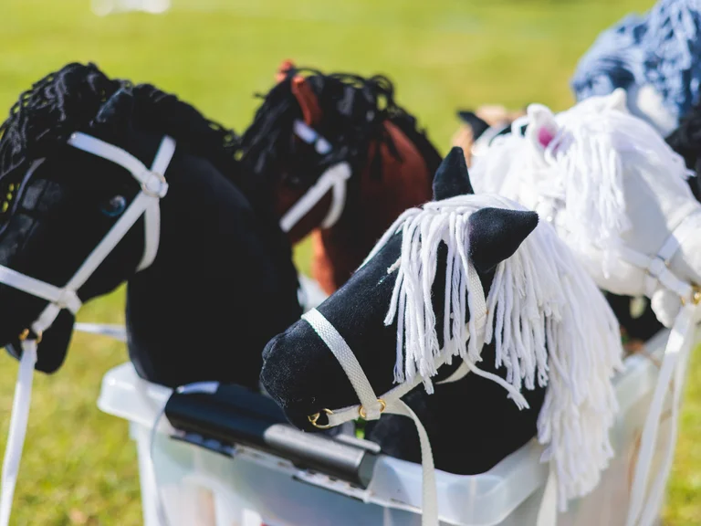 Several hobby horses with different colors and yarn manes stand side by side in a plastic box on a grassy field.