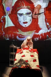 Scene from Balkan Erotic Epic: A person in traditional clothing paints the face of someone lying on a bed, while a large projection of a decorated face appears behind them.